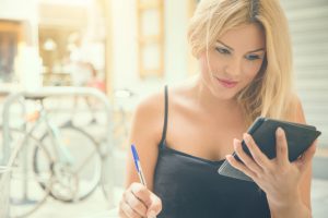 A young and beautiful woman, sitting by an outdoor table at a bar, study a book on her digital e-book reader, and write down notes on her agenda.